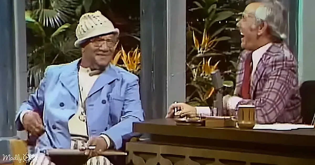 A man in a blue suit and hat is seated, holding a cigarette, while another man in a plaid suit sits behind a desk, laughing.
