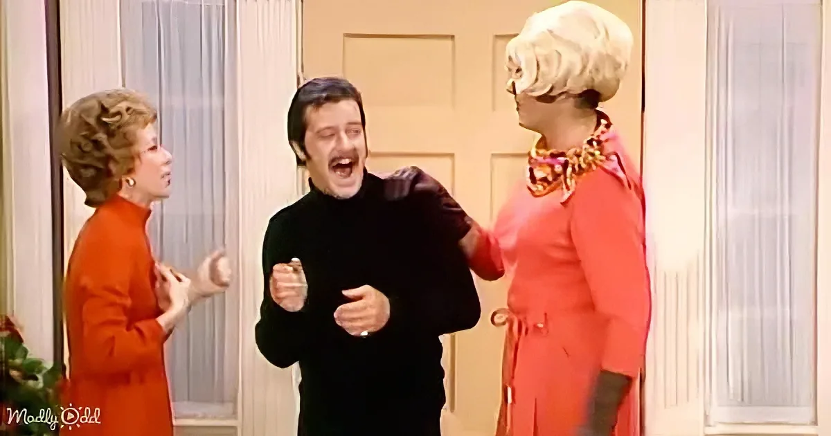 A man in a black turtleneck is excitedly gesturing while two women in red dresses look on.