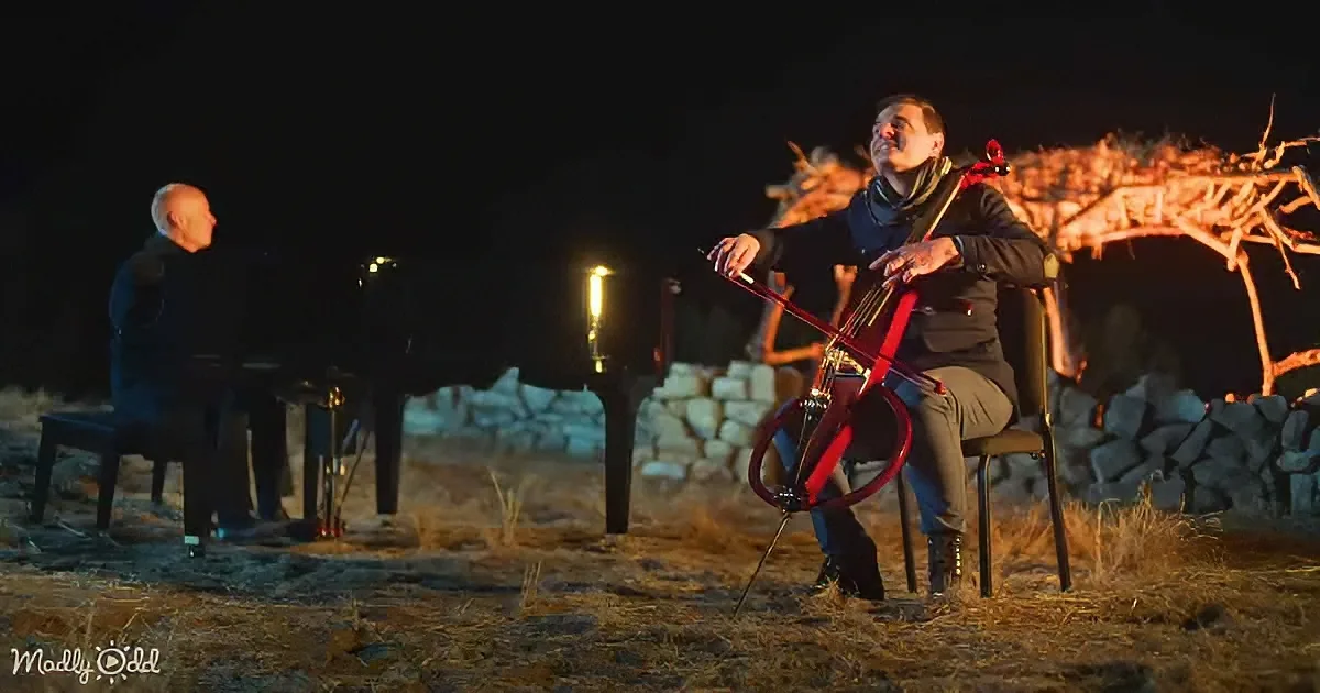 A man plays a red cello under a night sky, illuminated by a lantern, while another man plays a piano nearby.