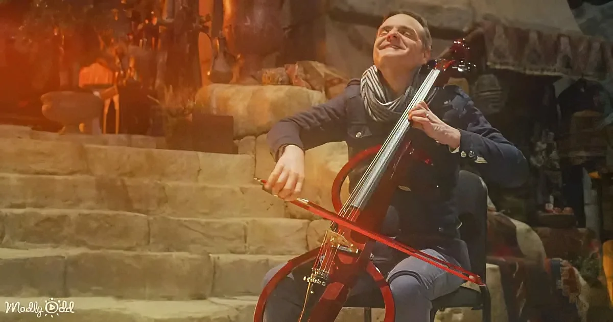 A man sits on stone steps, playing a red cello with a warm, glowing background.