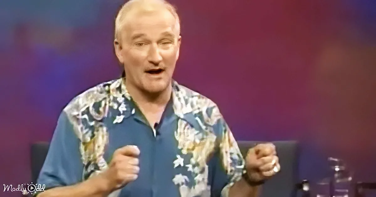 A man in a floral shirt is seated, gesturing with his fists, and appears to be speaking or reacting to something.
