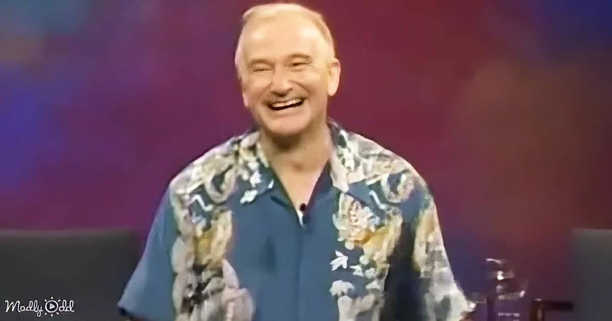 A man in a floral shirt smiles widely, seated against a colorful background.