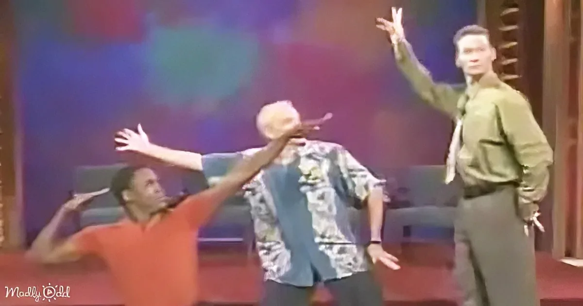 A man in a red shirt is dancing with his arms raised, while another man in a green shirt and tie stands nearby, also...