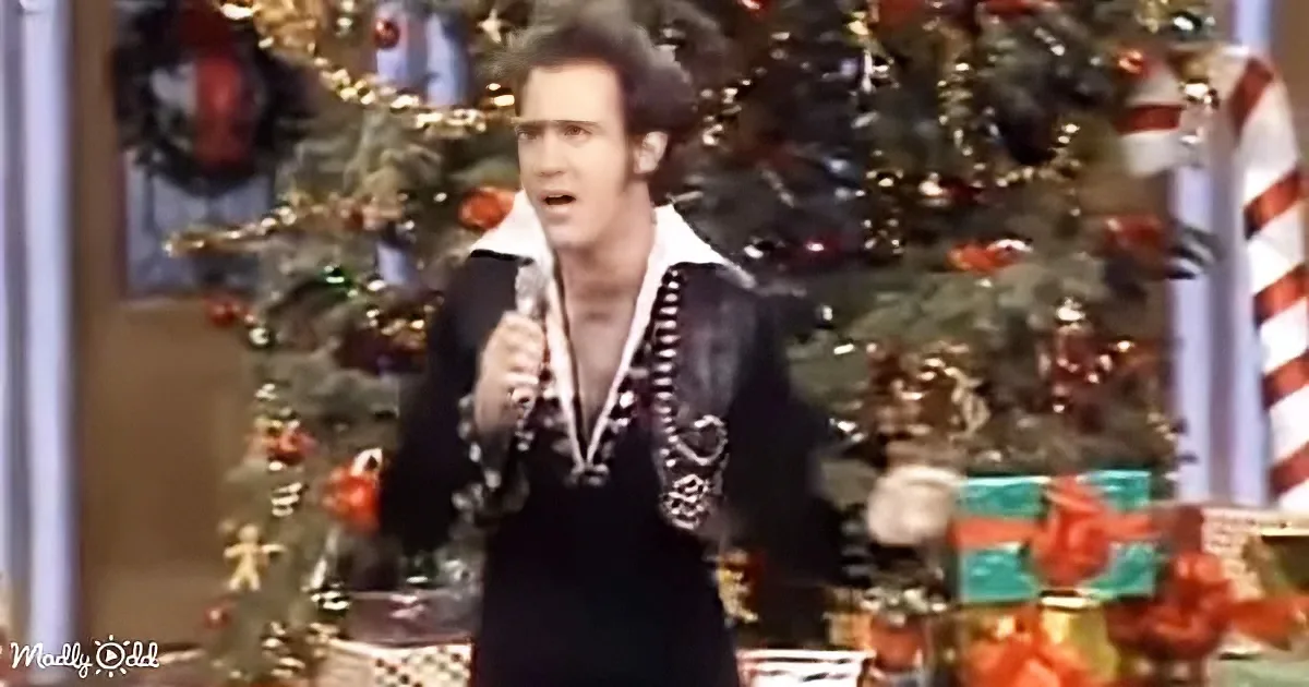 A person in a black outfit with white trim holds a microphone, standing in front of a Christmas tree and presents.