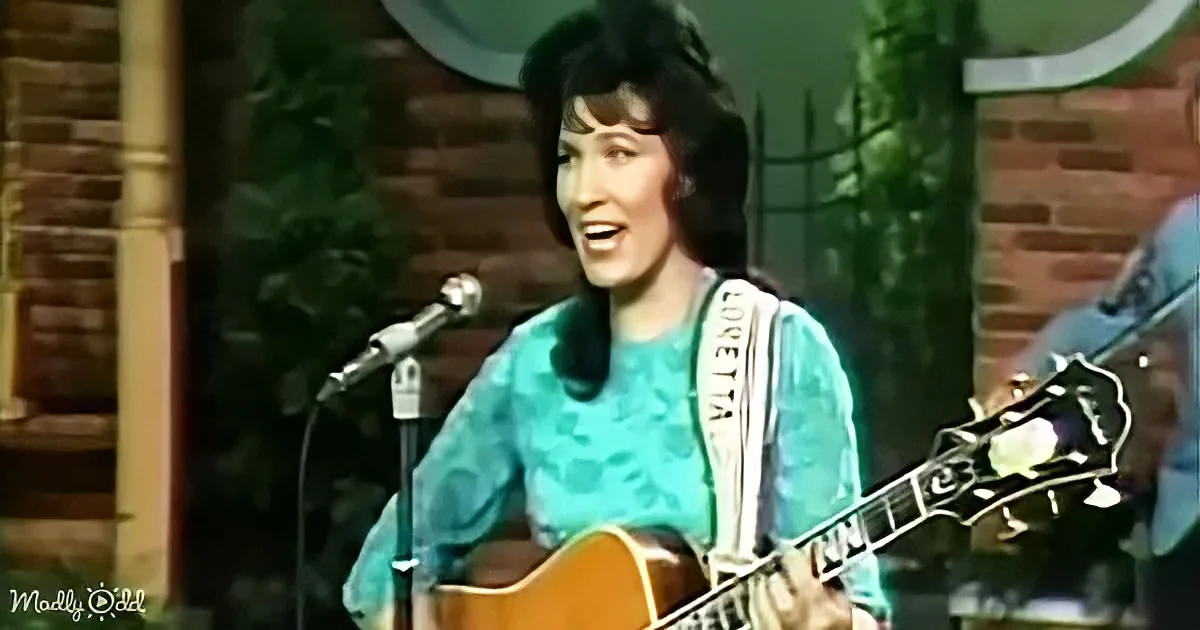 A woman in a teal dress plays guitar and sings on stage, with a brick wall and greenery in the background.