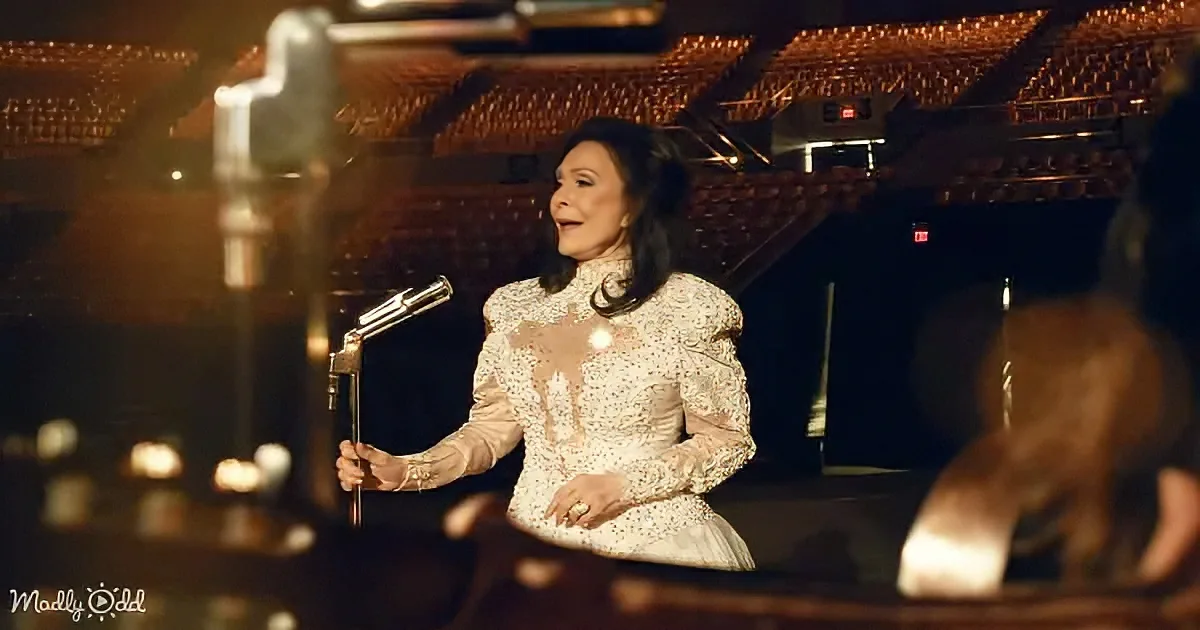 A woman in a white lace dress stands on stage, holding a microphone, with an empty auditorium in the background.