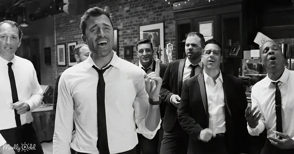 A man in a white shirt and black tie is smiling and pointing upwards, surrounded by other men in formal attire.