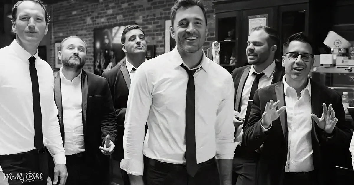 A man in a white shirt and black tie smiles, surrounded by five other men in formal attire.