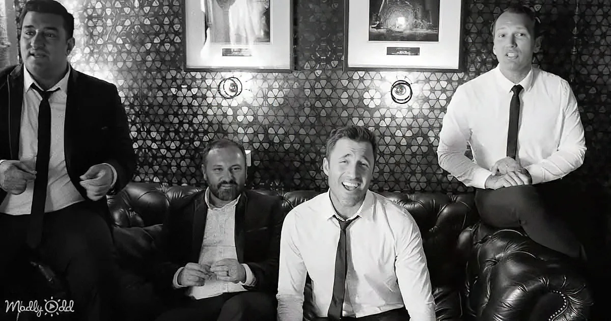 A man in a white shirt and tie sits on a leather couch, smiling, with three others in formal attire behind him.