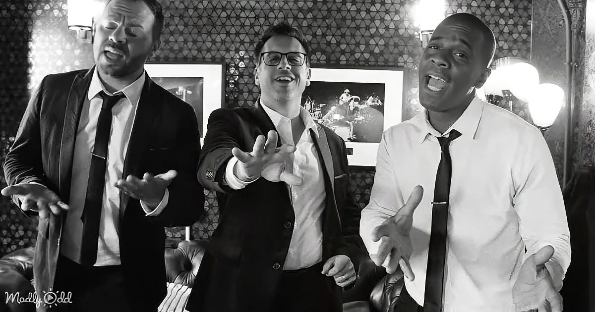 Three men in suits gesture dramatically in a dimly lit room with patterned walls.
