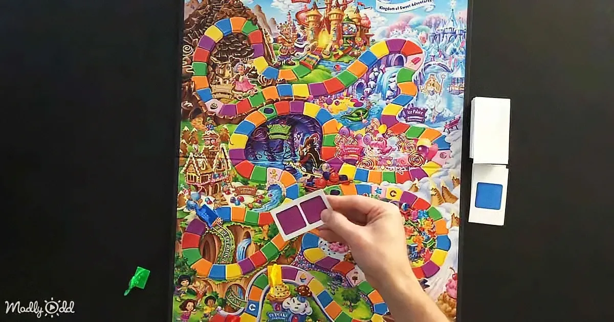 A hand holds a purple game piece over a colorful board game featuring a whimsical candy-themed landscape.