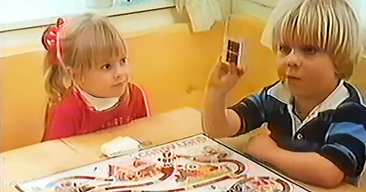 A young boy holds a small object while a girl watches, both seated at a table with a colorful game board.