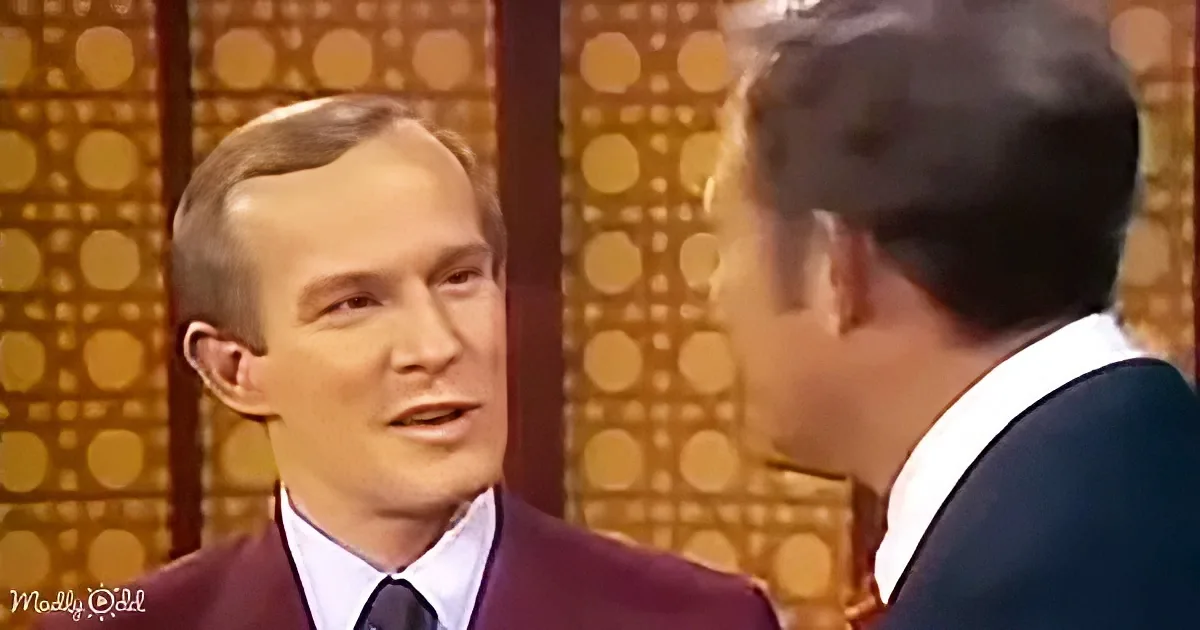 A man in a maroon suit and tie is speaking to another man in a dark suit against a patterned backdrop.