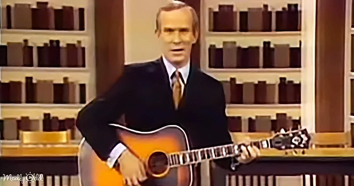 A man in a suit holds an acoustic guitar, standing in front of shelves filled with books.