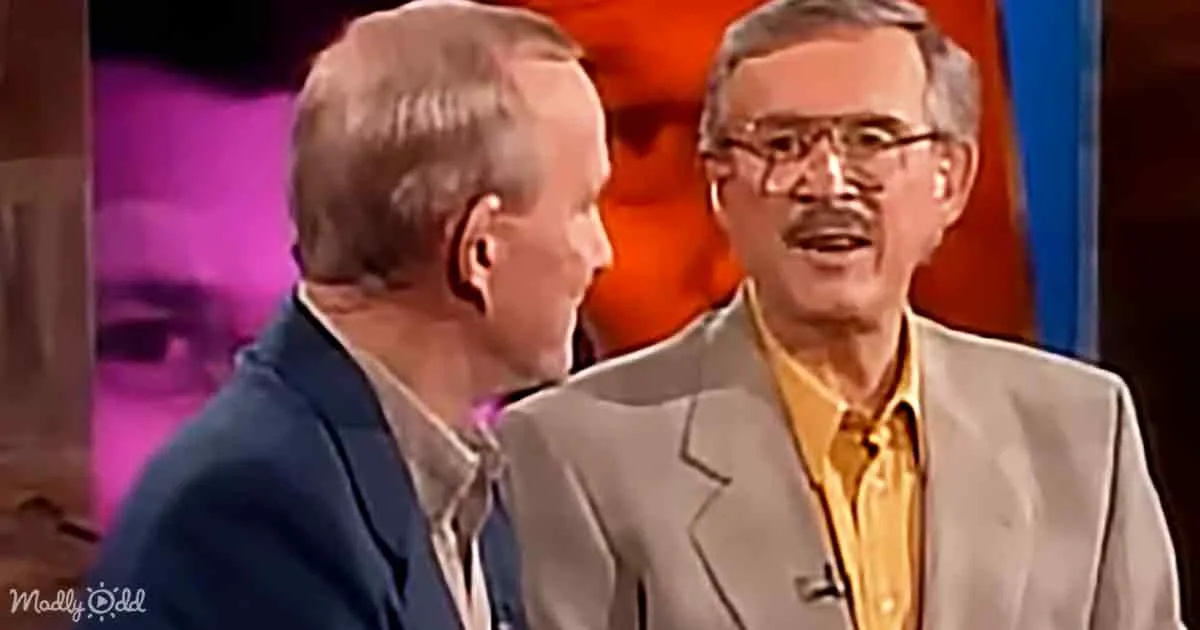 Two men in suits, one in a blue suit and the other in a beige suit with a yellow shirt, appear to be engaged in a...