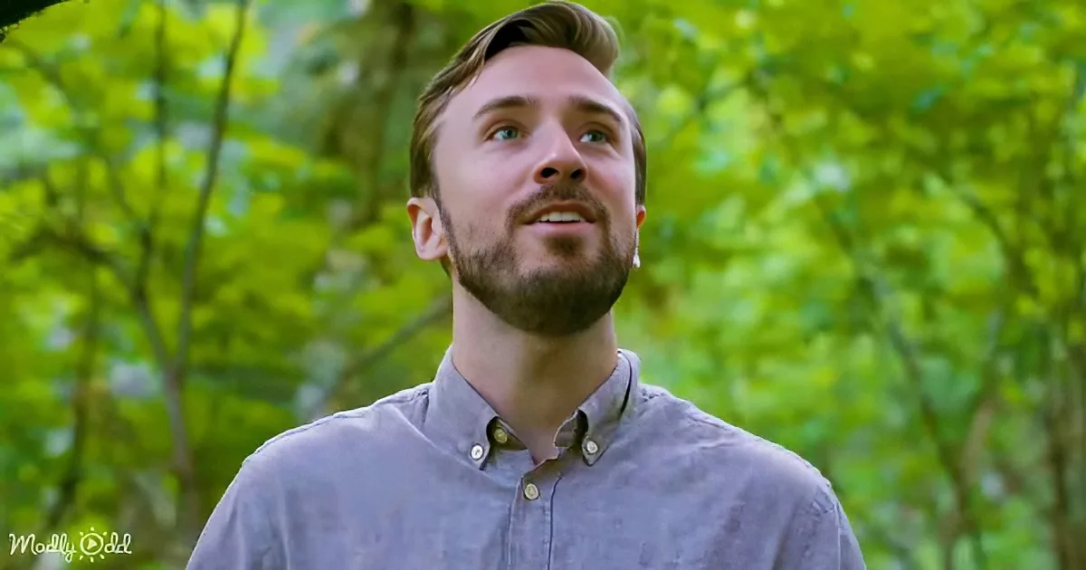 A man with a beard and short hair, wearing a gray shirt, looks up in a forest setting with green foliage.