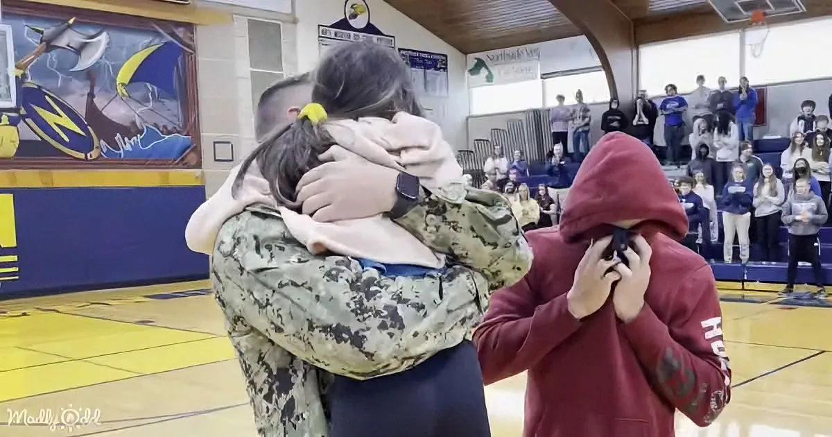 A soldier in camouflage embraces a child in a gymnasium, while another person covers their face nearby.