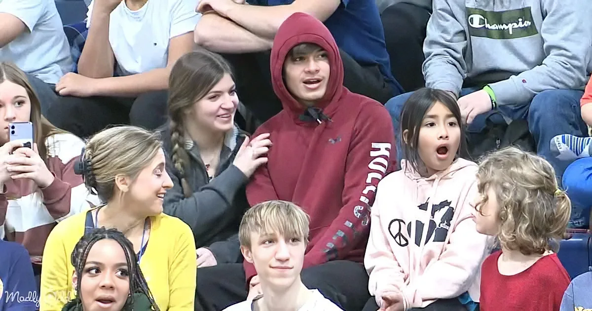 A young man in a red hoodie is surrounded by a group of people in a stadium setting, with varied expressions and attire.