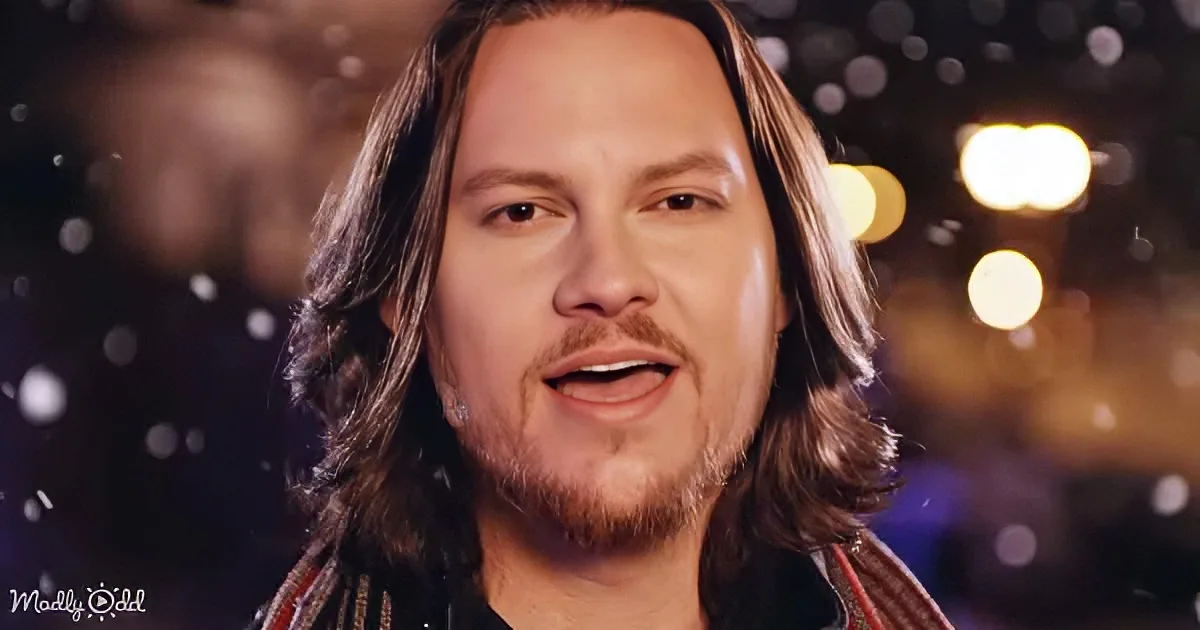 A man with long hair and a beard, wearing a dark shirt, smiles against a blurred background with bokeh lights.