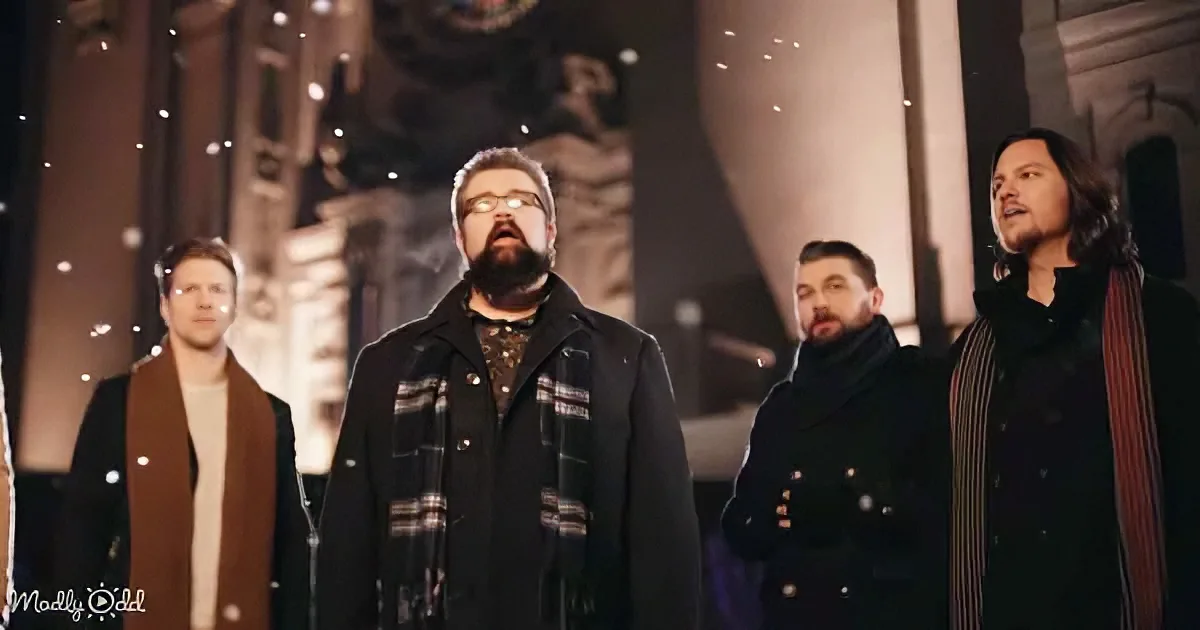 A man in a plaid scarf stands in front of a group of men, all dressed in coats, singing in a dimly lit setting.