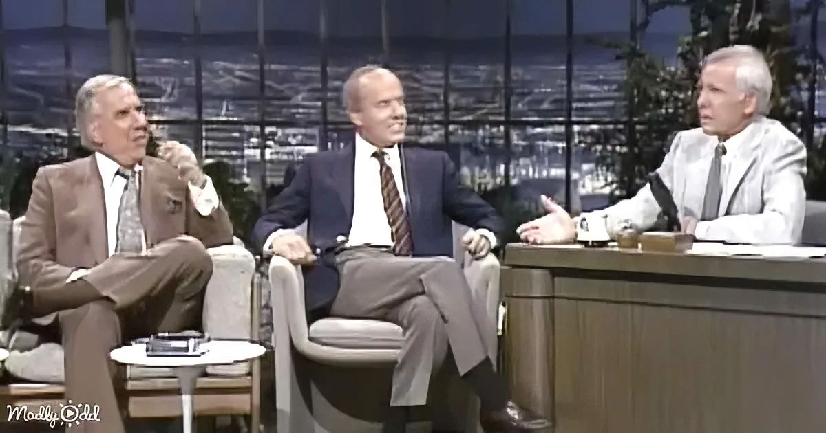 Three men in suits sit on a talk show set, engaged in conversation.