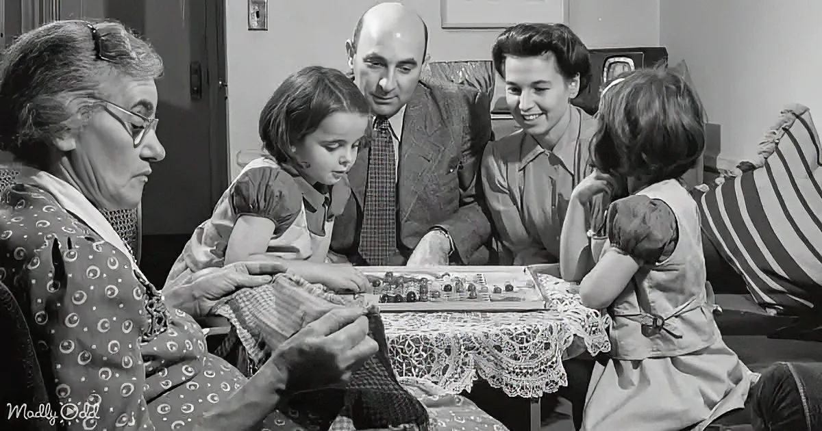 A family gathers around a table, playing a board game together, with a woman in glasses on the left.