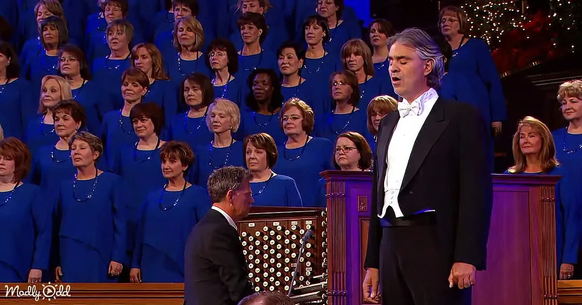 A man in a black suit sings in front of a choir in blue robes, with an organist seated at a pipe organ.