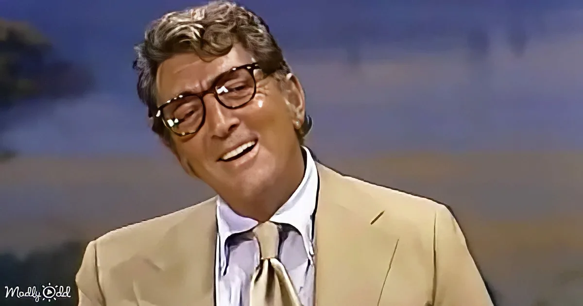 A man in a beige suit and glasses smiles, wearing a light blue shirt and beige tie, against a blurred background.