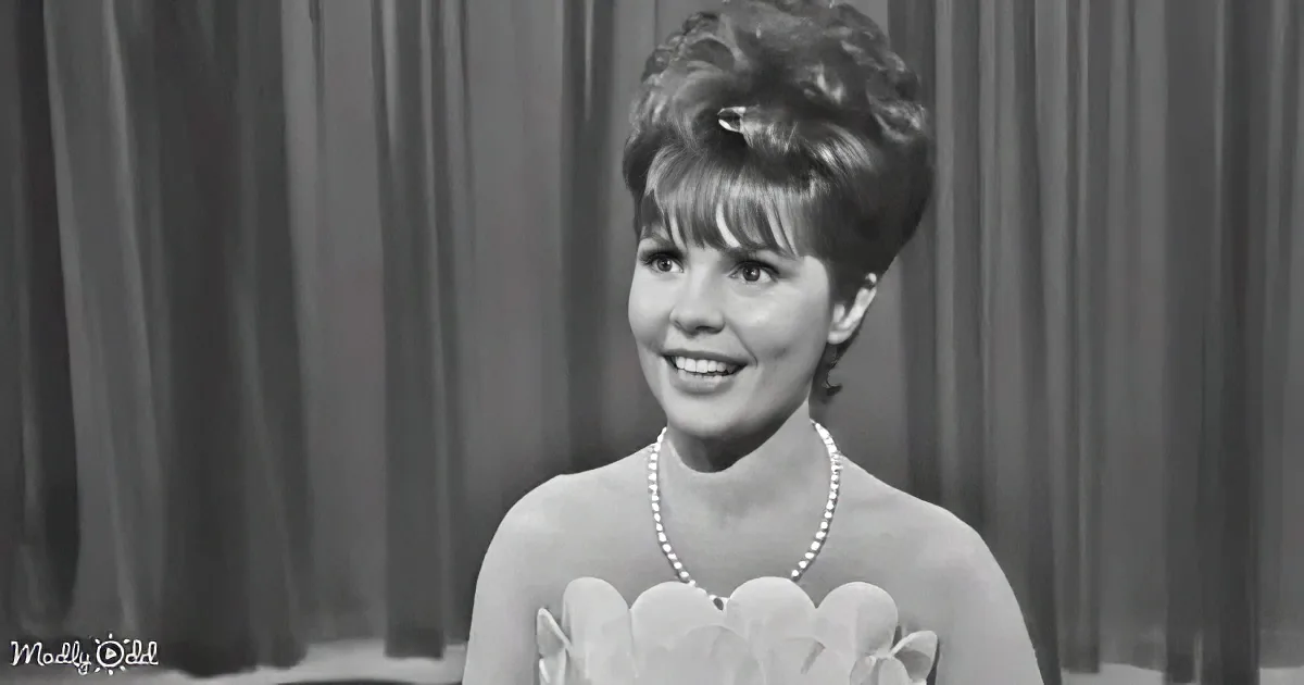 A woman in a floral dress and pearl necklace smiles against a curtain backdrop.
