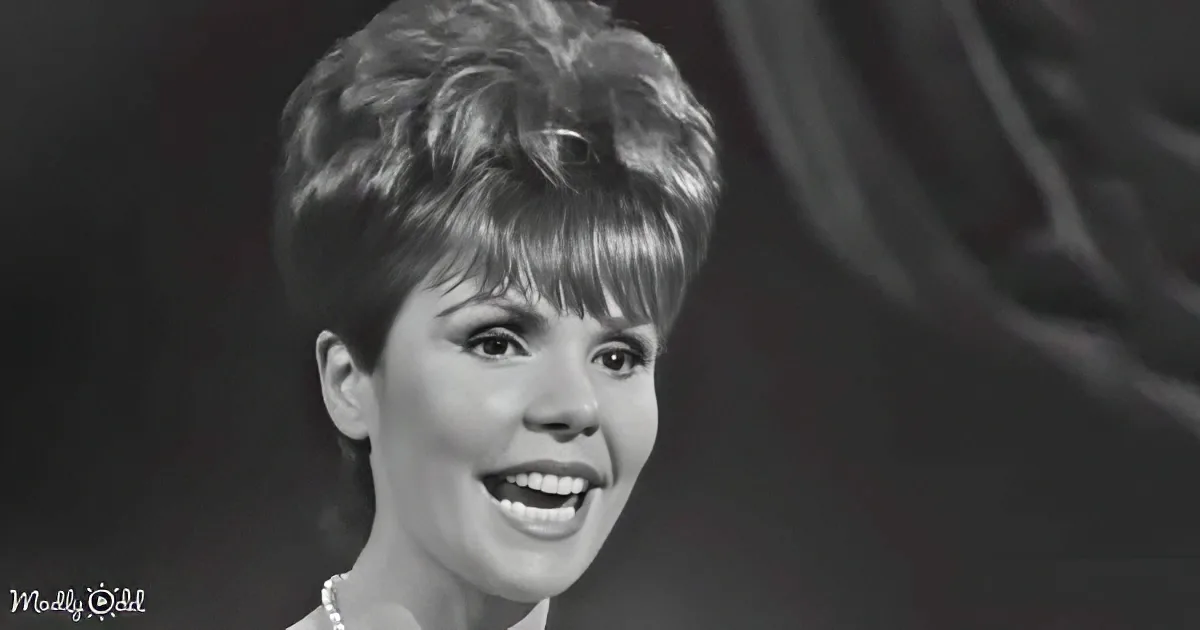 A woman with short, styled hair and a pearl necklace smiles warmly, set against a dark background.