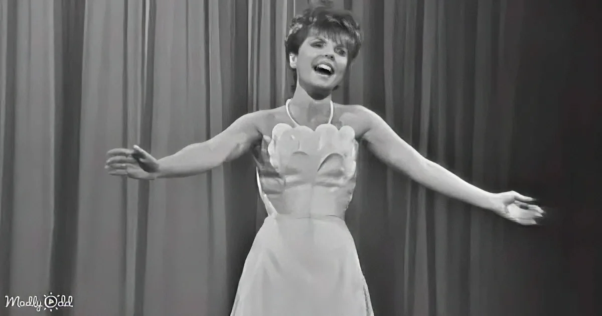 A woman in a white dress with a heart-shaped neckline is singing with arms outstretched.