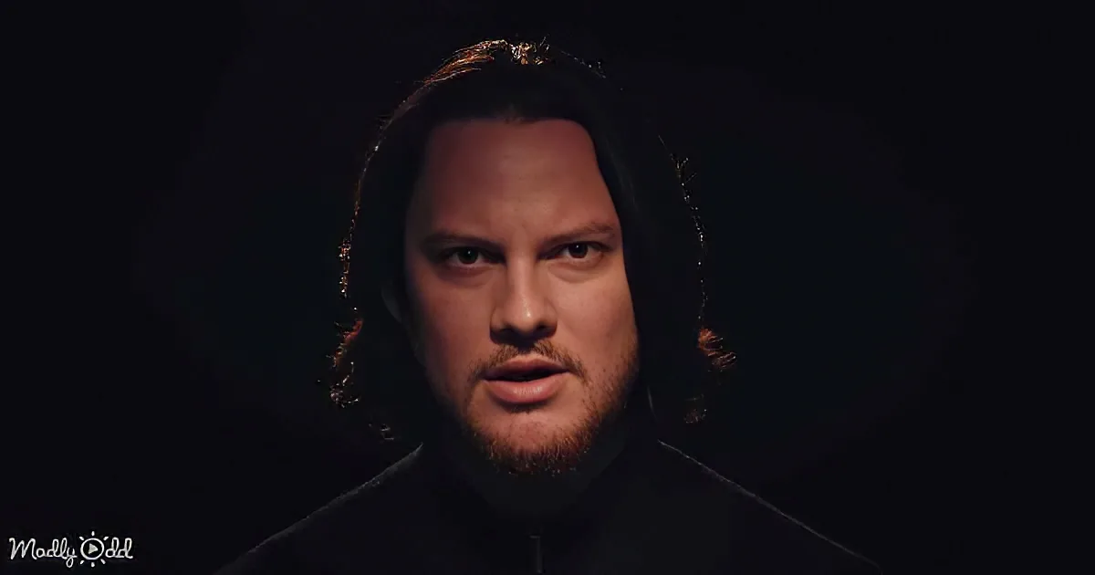 A man with long hair and a beard, wearing a dark hoodie, stands against a dark background.