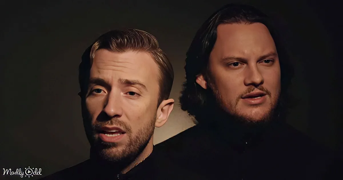 Two men with beards, one with short hair and the other with long hair, both wearing dark clothing, against a dark background.
