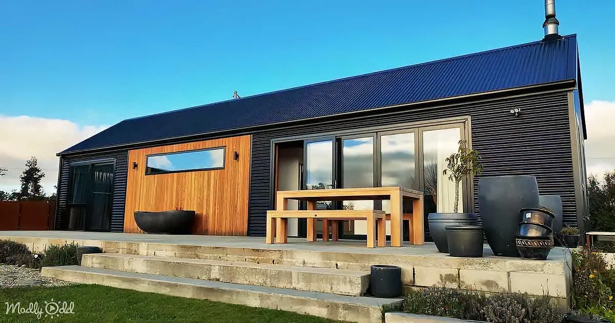 A modern house with a dark blue roof, wooden accents, and a patio with a table and potted plants.