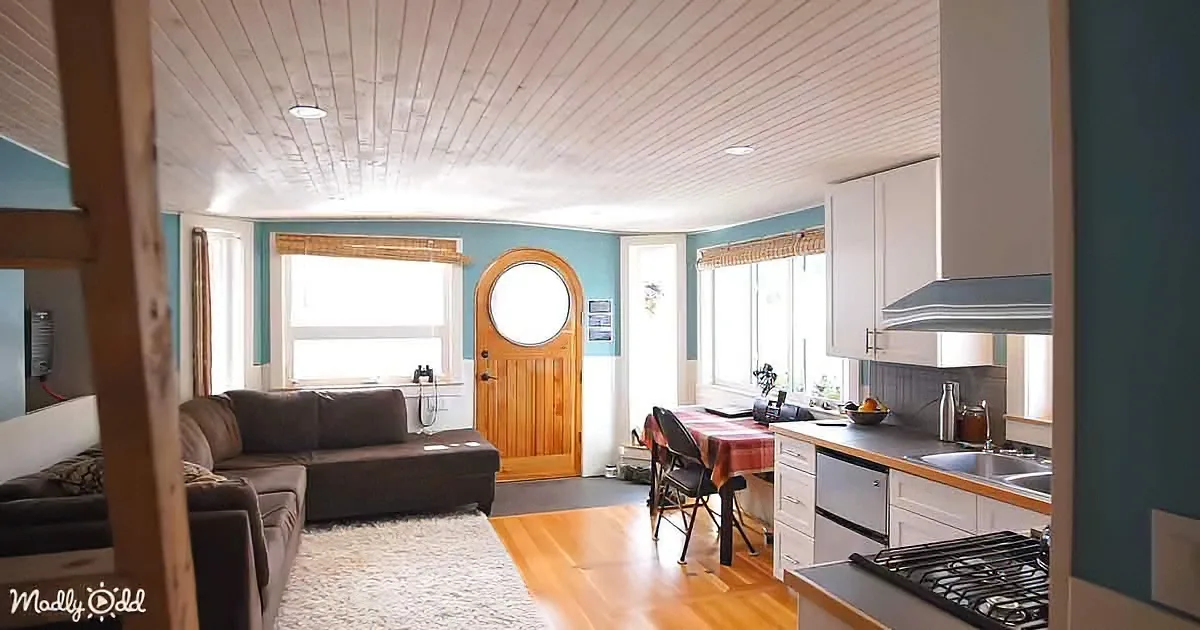 A cozy living room with a sectional sofa, wooden floor, and kitchen area with white cabinets and a round window.