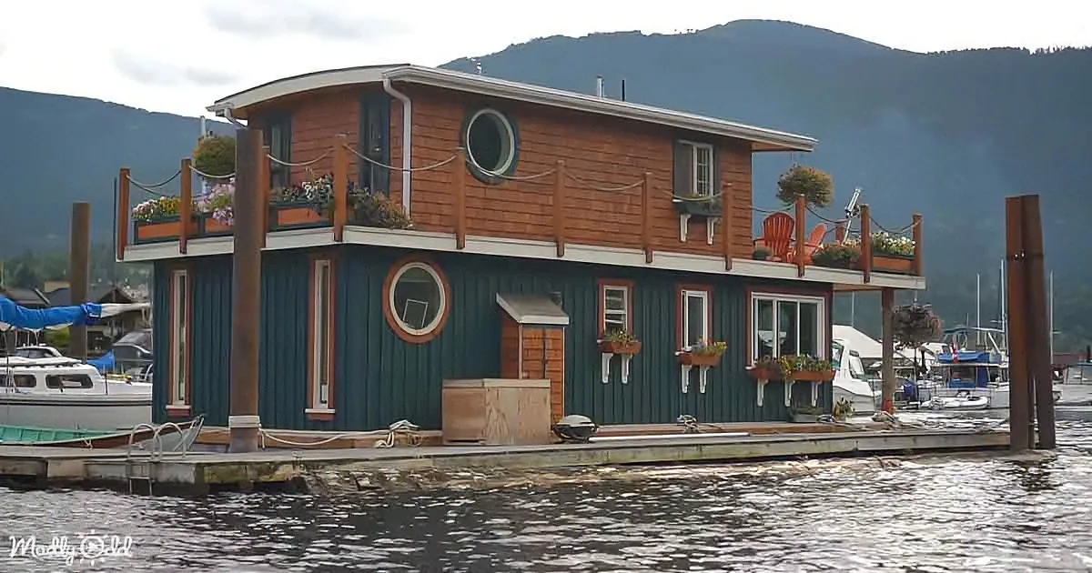 A houseboat with a green and brown exterior, adorned with flowers, sits on calm water with mountains in the background.