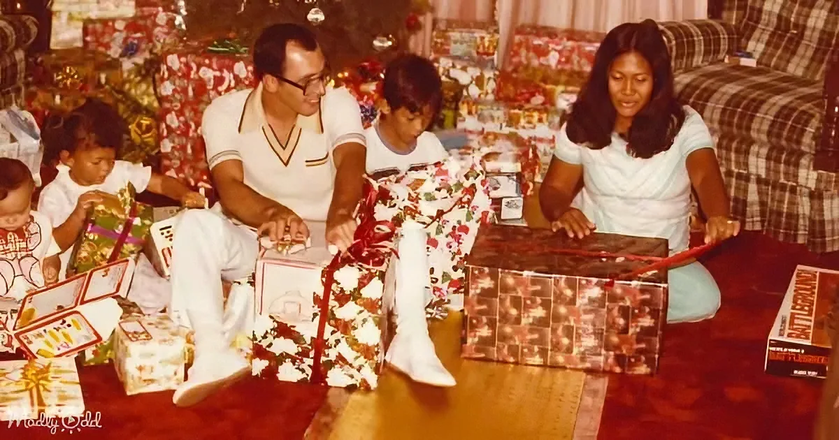 A family of five unwraps Christmas presents on the floor, surrounded by festive decorations and gifts.