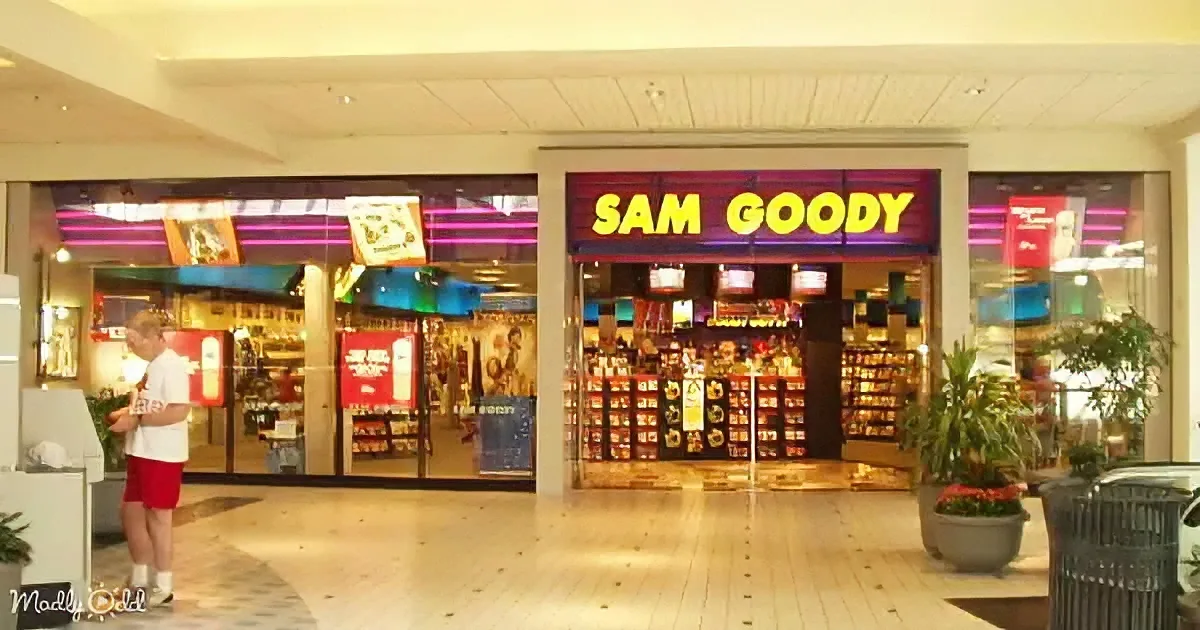 Man in red shorts stands outside Sam Goody store in mall with bright neon lights.