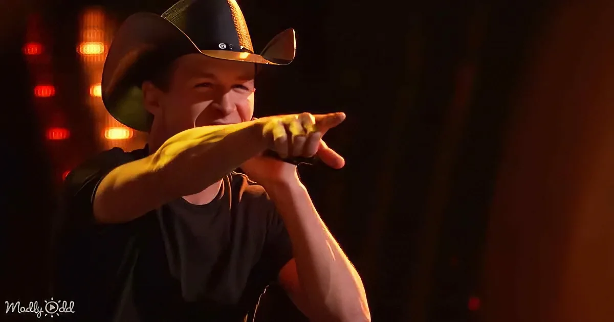 A person in a cowboy hat points forward with a microphone, illuminated by stage lights.