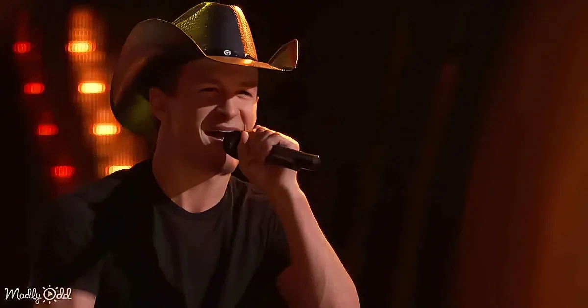 A person in a cowboy hat sings into a microphone, illuminated by stage lights.