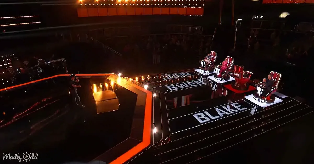 A stage with judges' chairs labeled John, Gwen, and Blake, illuminated by stage lights.