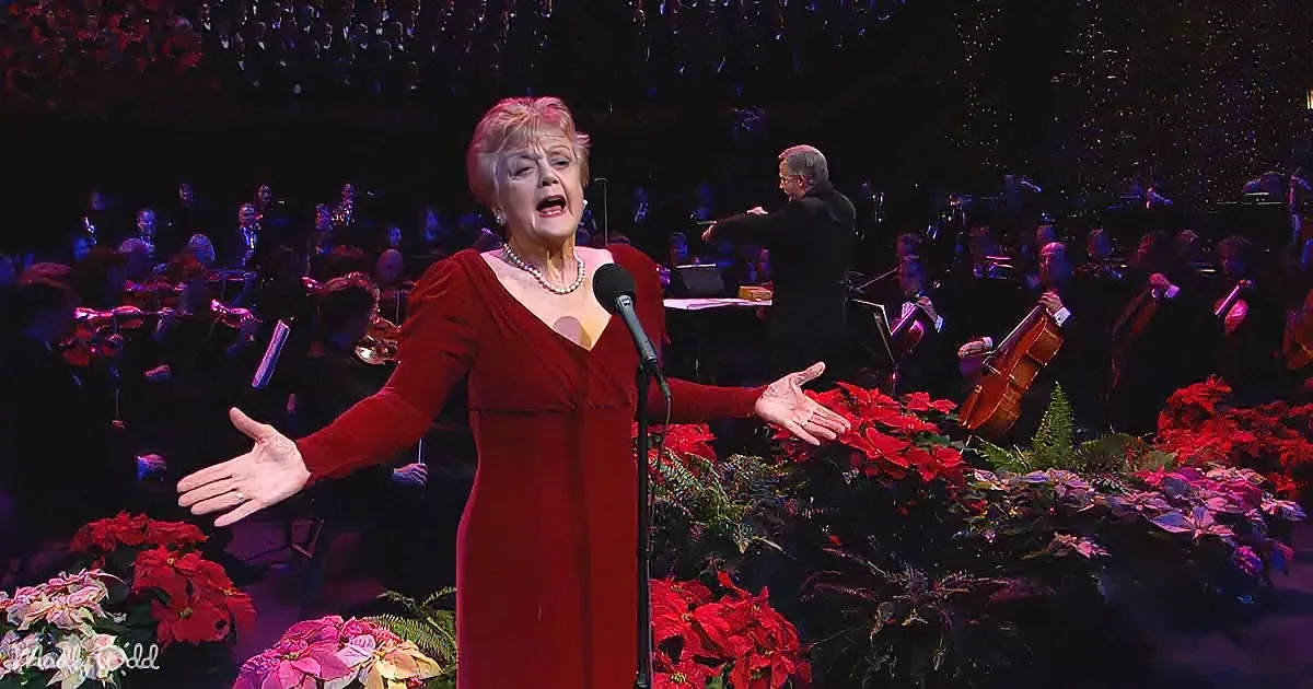 A woman in a red dress sings on stage with an orchestra and poinsettias in the foreground.