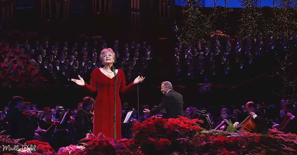 A woman in a red dress sings on stage with a choir and orchestra behind her.