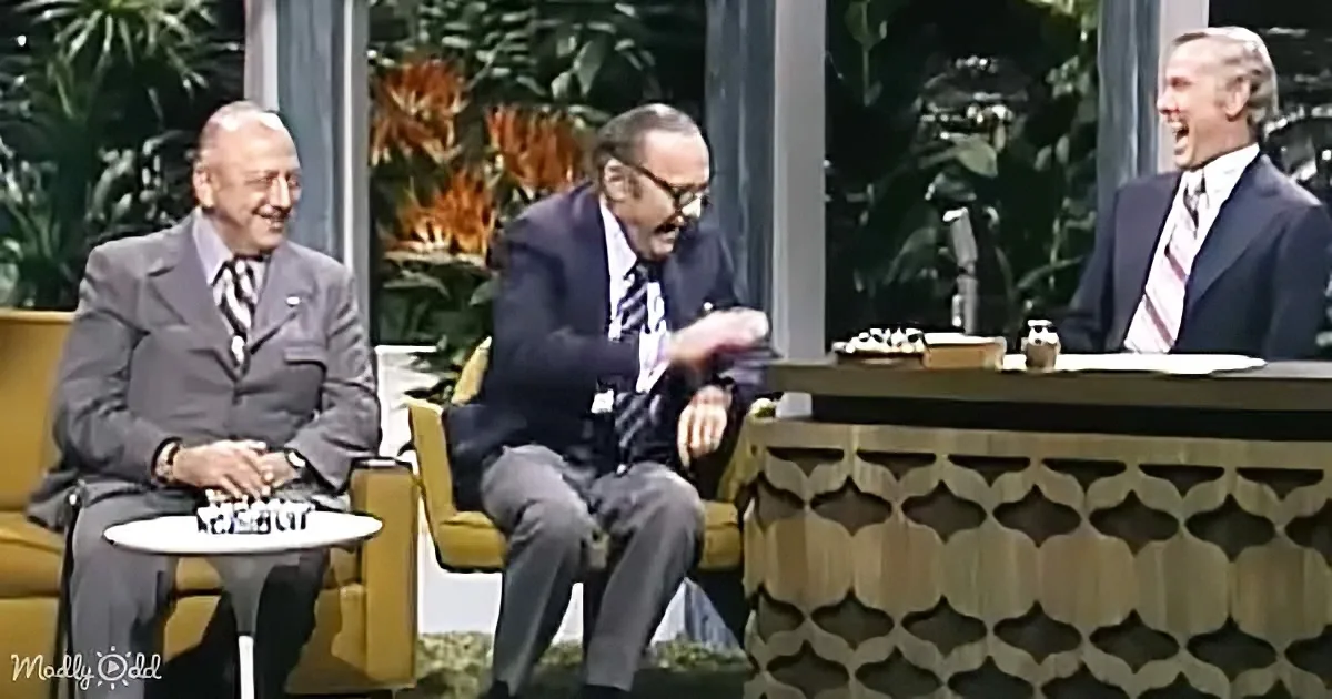 Three men in suits, one laughing, on a talk show set with a patterned desk and plants in the background.