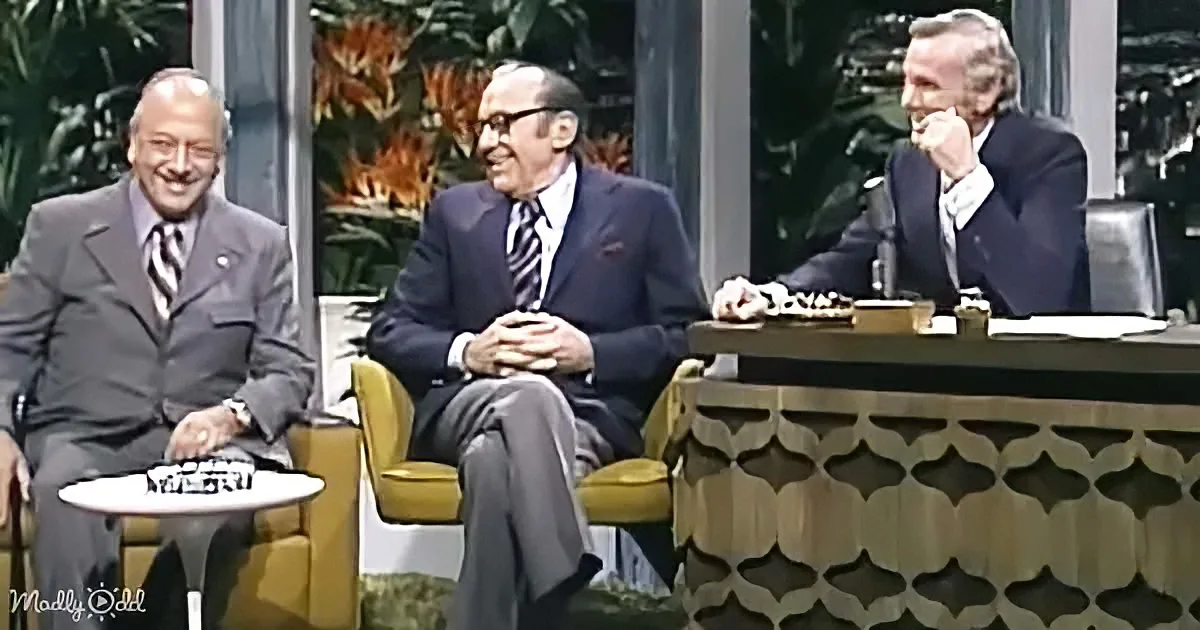 Three men in suits sit on a talk show set, one gesturing, another smiling, and the third appearing thoughtful.