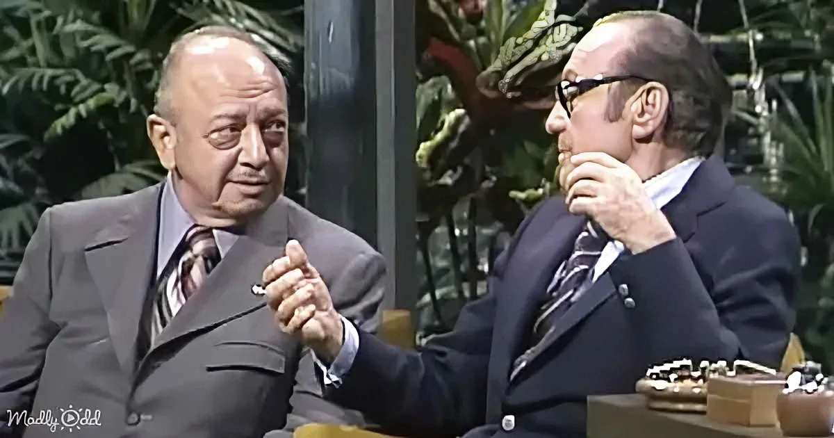 Two men in suits are seated, one gesturing while the other listens, with a backdrop of greenery.