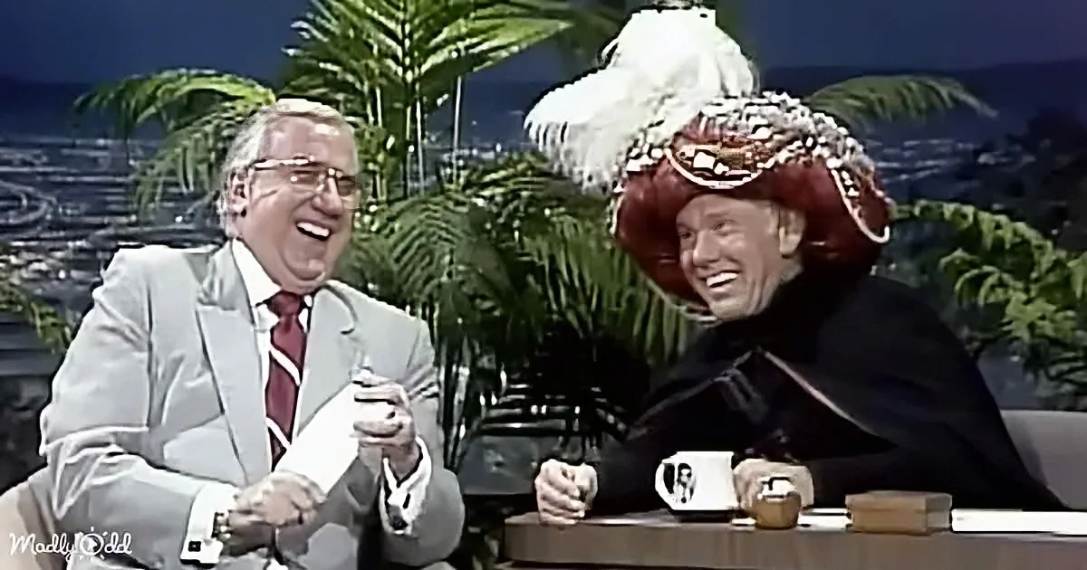 Two men in suits laugh, one in a red hat with feathers, on a talk show set with palm trees.