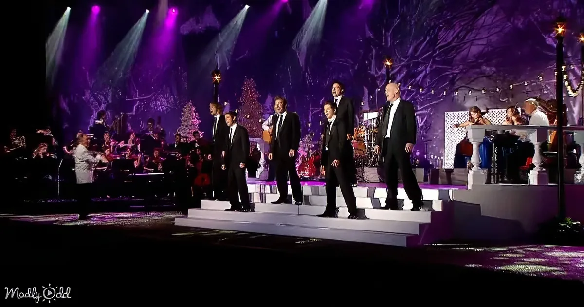A group of men in black suits sing on stage with a band and Christmas decorations behind them.