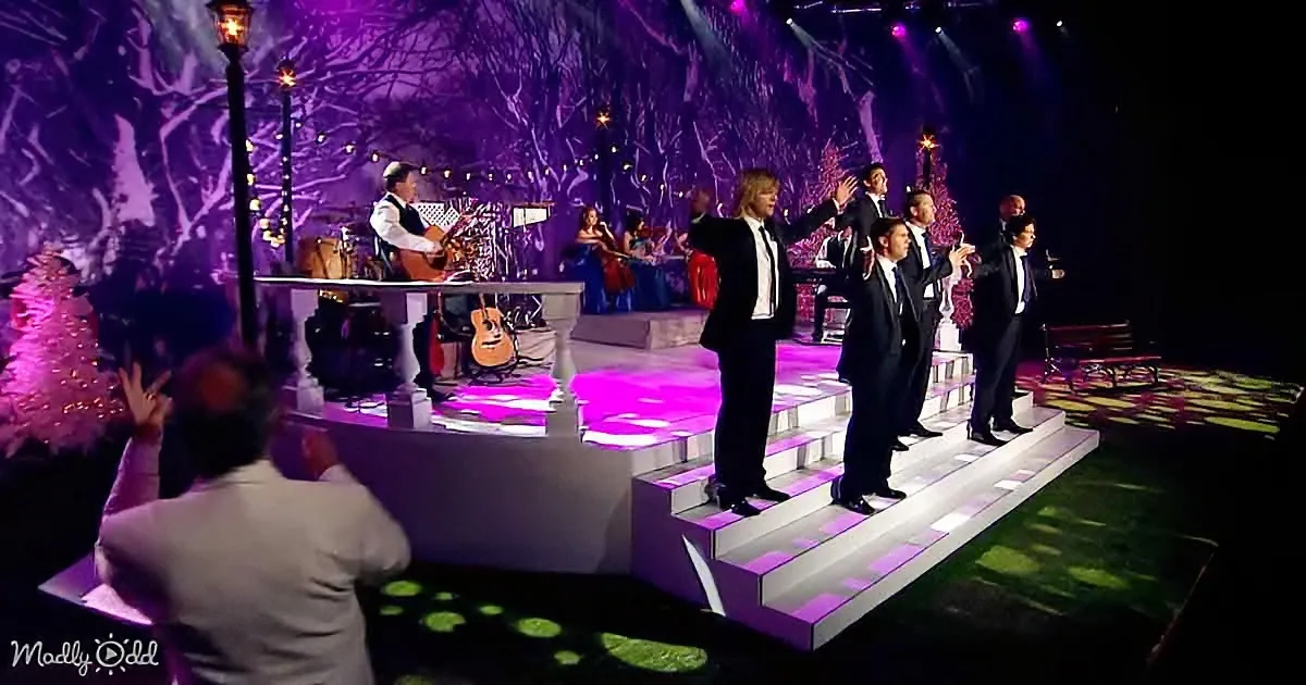 A group of men in suits stands on steps, singing, with a band and Christmas tree in the background.