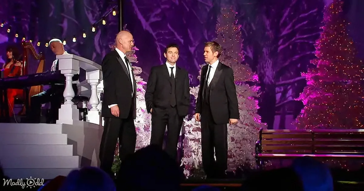 Three men in suits stand on stage, surrounded by Christmas decorations and a performer.